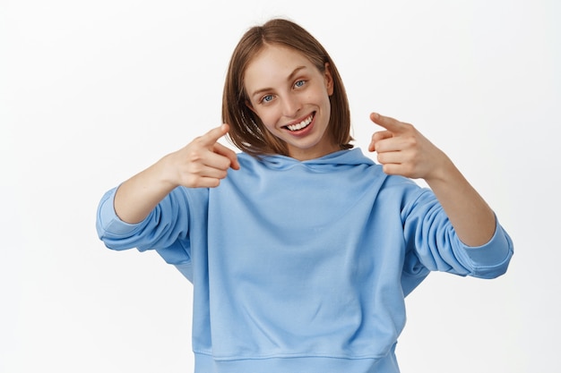 Smiling blond woman pointing fingers at front with happy proud face, praise and compliment, invite people, beckon to join company, standing against white wall.