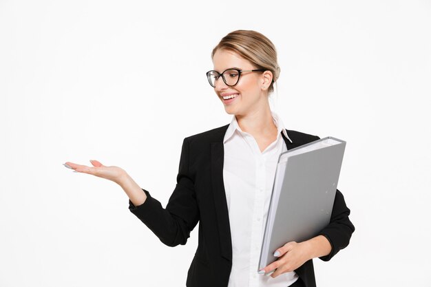 Smiling blonde business woman in eyeglasses with folder in hand holding copyspace on the pound over white wall