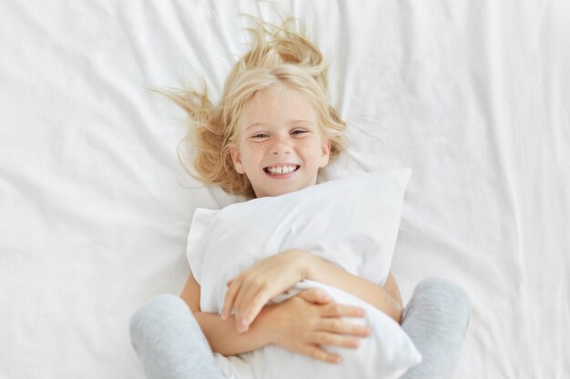 Smiling blonde girl embracing white pillow while being in kindergarten, having good mood while seeing someone and lying in white bed. Little adorable female child having beddtime. Rest concept