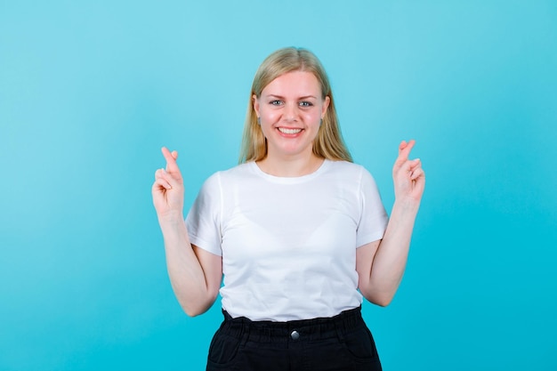 Smiling blonde girl is looking at camera by crossing fingers on blue background