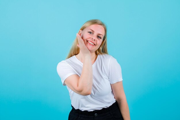 Smiling blonde girl is looking at camera by holding hand under chin on blue background