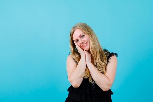 Smiling blonde girl is looking at camera by holding handfuls together on cheek on blue background