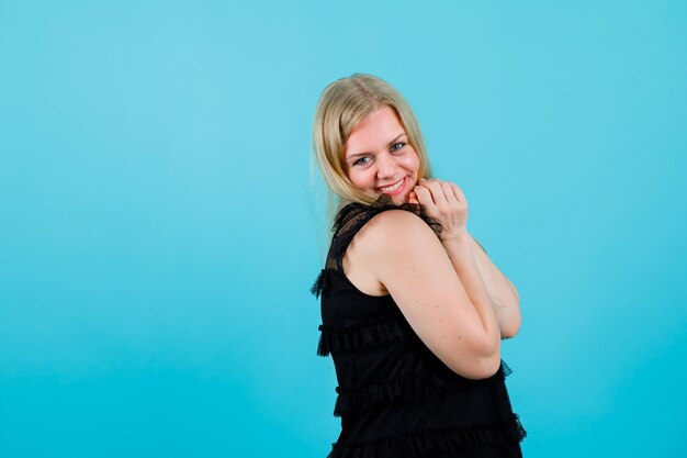 Smiling blonde girl is looking at camera by holding hands on chest on blue background