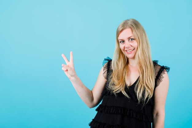 Smiling blonde girl is looking at camera by hsowing victory gesture on blue background