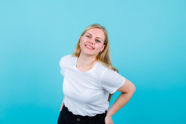 Smiling blonde girl is looking at camera by putting hand on waist on blue background