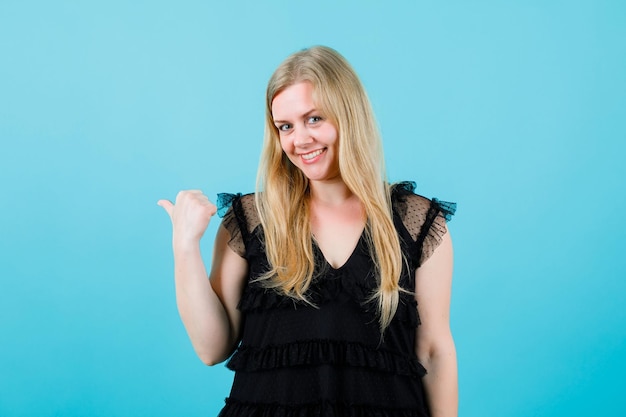 Smiling blonde girl is pointing back with thumb on blue background