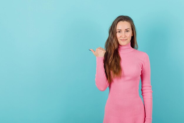 Smiling blonde girl is pointing to left with thumb on blue background