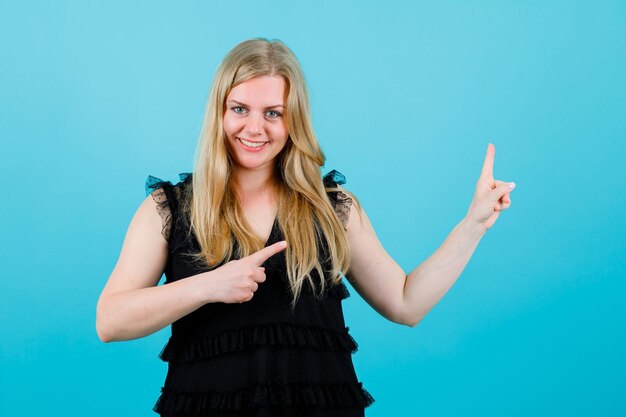 Smiling blonde girl is pointing right up with forefingers on blue background