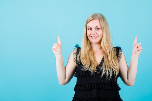 Smiling blonde girl is pointing up with forefingers on blue background