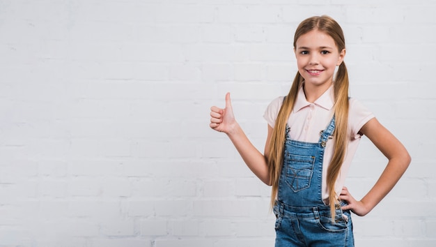 Ragazza bionda sorridente che mostra pollice sul segno che sta contro il muro di mattoni bianco