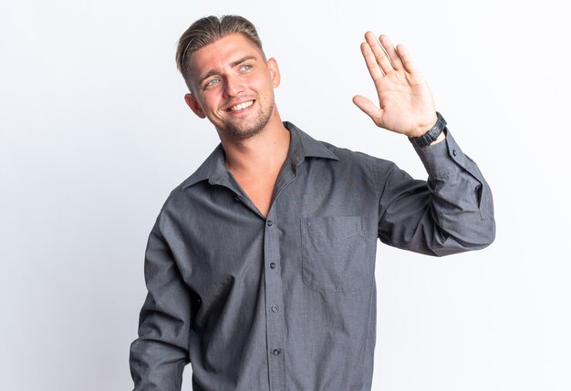 Smiling blonde handsome man standing with raised hand and looking at side isolated on white wall with copy space