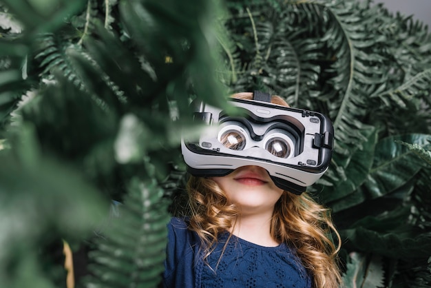 Smiling blonde little girl using virtual headset standing among the green plant