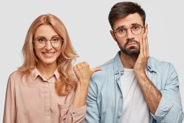 Smiling blonde woman model indicates with thumb at displeased brother who feels puzzled