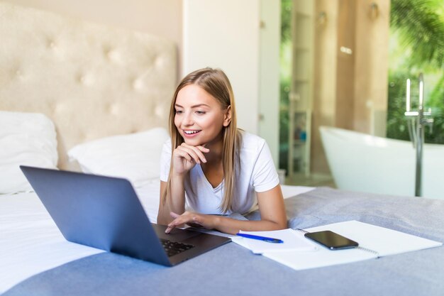 Smiling blonde woman using laptop at home in bedroom