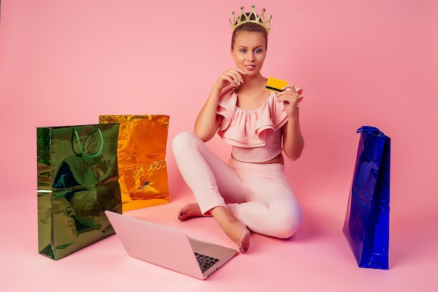 Smiling blonde woman with a crown on head shopaholic bag and credit card in her hand sitting on floor with laptop pink background in studio . concept of seasonal black friday sale and online shopping.