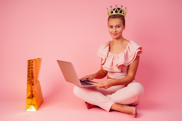 Smiling blonde woman with a crown on head shopaholic bag and credit card in her hand sitting on floor with laptop pink background in studio . concept of seasonal black friday sale and online shopping