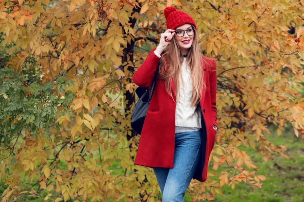 Smiling blonde woman with long hairs walking in sunny autumn park in trendy casual outfit.