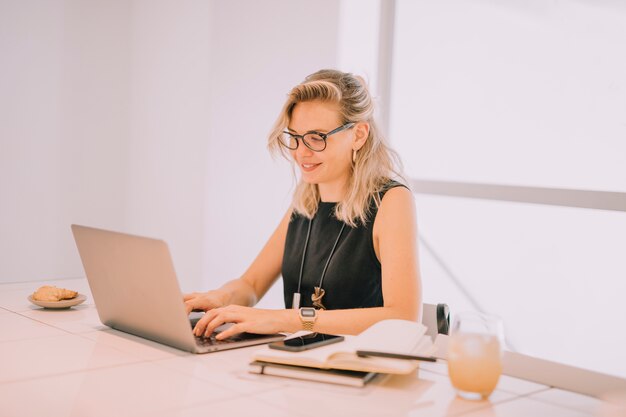 Smiling blonde young businesswoman using laptop with breakfast on office table