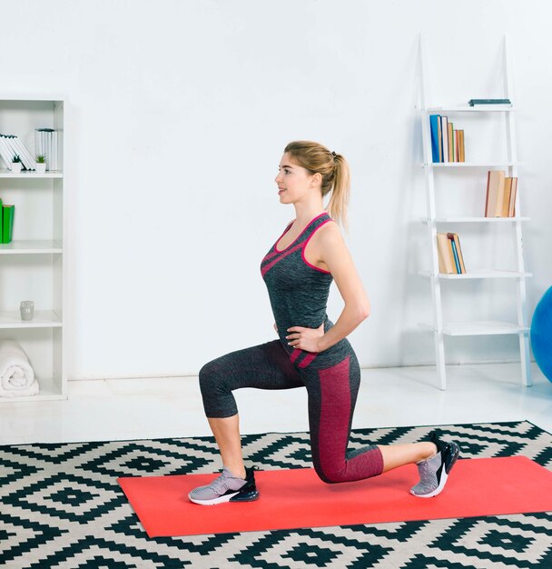 Smiling blonde young woman exercising on red mat at home in the living room