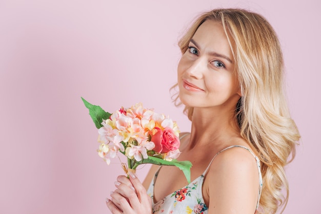 Smiling blonde young woman holding flower bouquet against pink backdrop