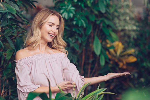 Smiling blonde young woman showing open hand palm standing near the plant