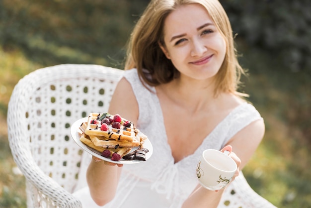 Giovane donna bionda sorridente che mostra piatto delle cialde e della tazza di caffè