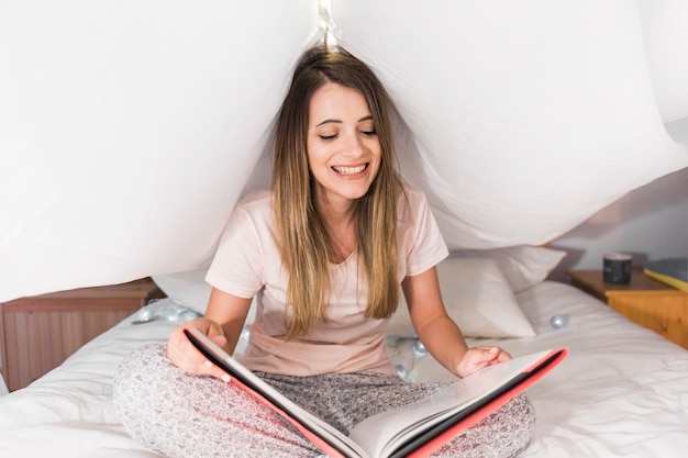 Free photo smiling blonde young woman sitting on bed reading magazine