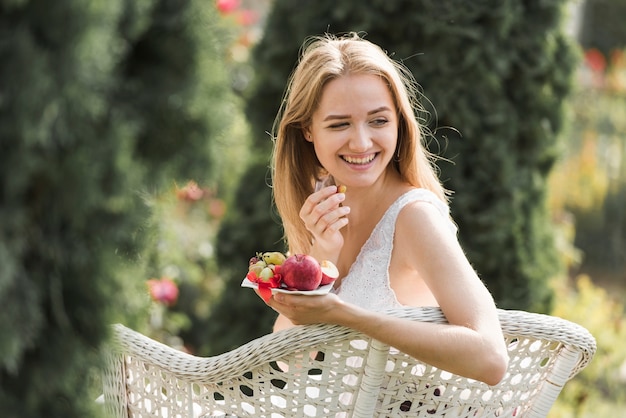 Улыбаясь блондинка молодая женщина, сидя на стуле, едят фрукты в саду