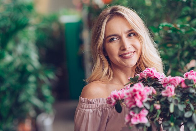Усмехаясь белокурая молодая женщина стоя перед цветковыми растениями