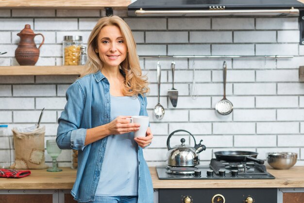 白いコーヒーカップを保持しているガスストーブの近くに立っている笑顔金髪の若い女性