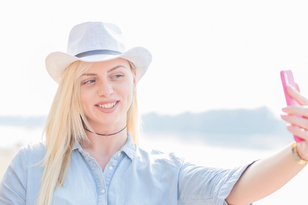 Giovane donna bionda sorridente che prende selfie dal telefono cellulare