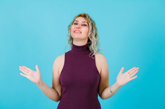 Smiling blondew oman is looking at camera by opening wide her hands on blue background