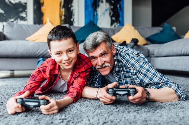 Ragazzo sorridente e suo nonno che giocano insieme ai videogiochi a casa, sdraiato sul pavimento.