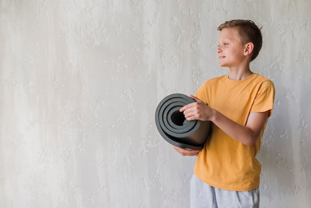 Smiling boy holding rolled up exercise mat looking away in front of concrete wall