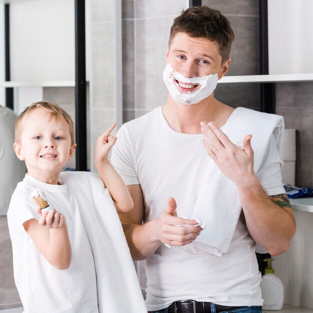 Smiling boy holding shaving brush in hand pointing finger toward his father with shaving foam on his face