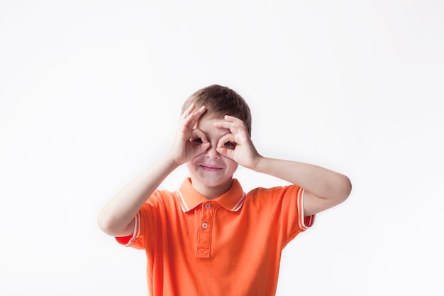 Smiling boy looking through ok hand gesture on white backdrop