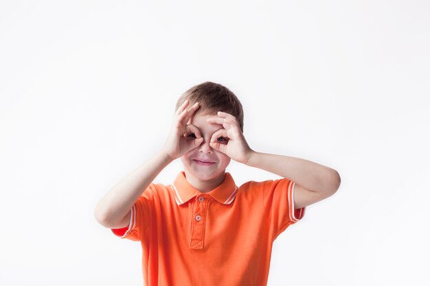 Smiling boy looking through ok hand gesture on white backdrop