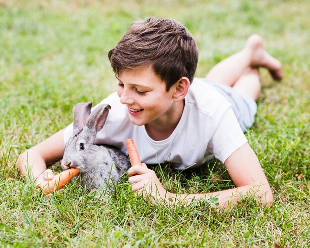 Smiling boy lying on green grass feeding carrot to rabbit