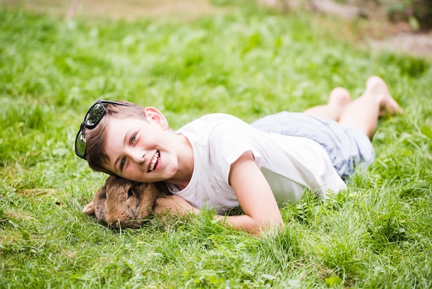 Smiling boy lying with rabbit on green grass in the park