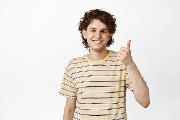 Улыбающийся мальчик показывает большой палец вверх и выглядит счастливым, стоя в футболке на белом фоне
