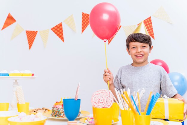 Smiling boy wearing party hat holding balloon and gift standing behind variety of food on table