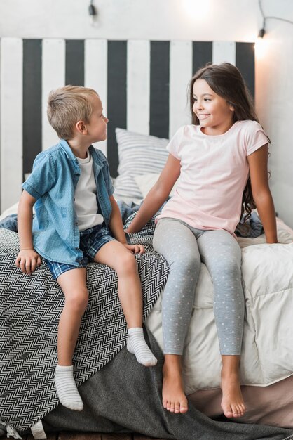 Smiling brother and girl sitting on bed looking at each other