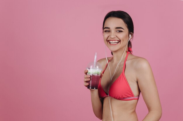 Smiling brunette in a bikini stands with earphones and cold cocktail
