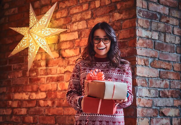 안경을 쓰고 따뜻한 스웨터를 입은 웃고 있는 갈색 머리 여성이 벽돌 벽에 크리스마스 선물을 들고 있습니다.