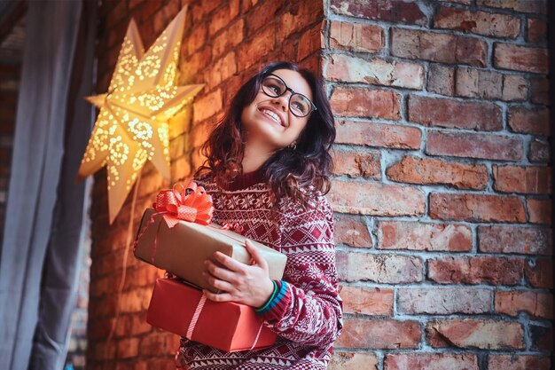 Smiling brunette female in eyeglasses and a warm sweater holds Christmas gifts over the wall of a brick.