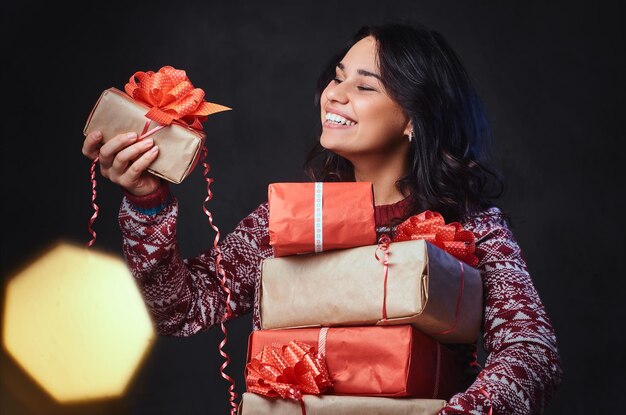 Smiling brunette female in eyeglasses and a warm sweater holds Christmas gifts with bright color effect on the image.