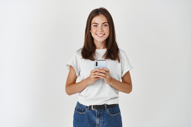 Foto gratuita sorridente donna bruna che tiene smartphone e guarda davanti, in piedi in una maglietta casual sul muro bianco.