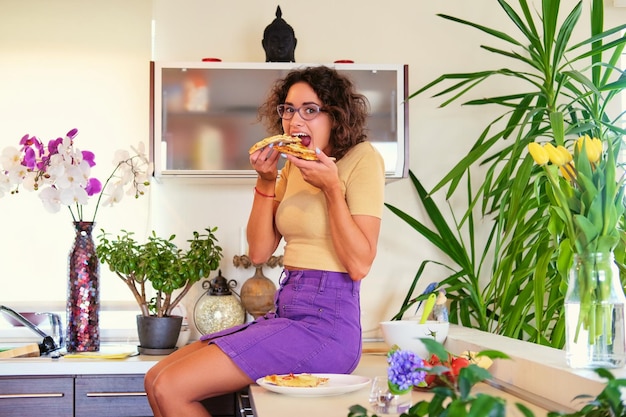 곱슬머리를 한 웃고 있는 브루네트 여성은 바닥에 앉아서 부엌에서 피자를 먹습니다.