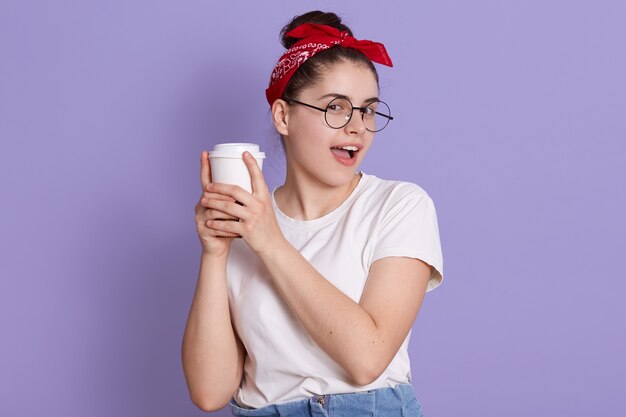 Smiling brunette girl in white casual t shirt holding cup of coffee isolated over lilac space, looking into at camera, keeps mouth opened