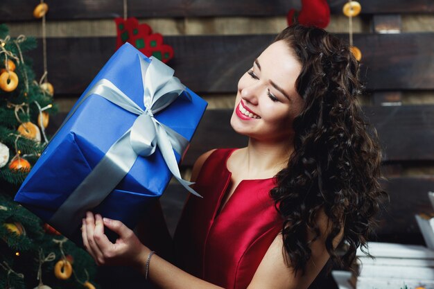 Smiling brunette holds blue present box standing before Christmas tree